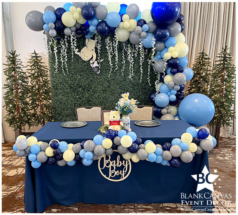 Winnie the Pooh Head Table Backdrop. Greenery backdrop rental with organic style balloon garland in yellow, light blue, grey and navy  with flowering vines hanging down and a Winnie the Pooh cutout with Piglet